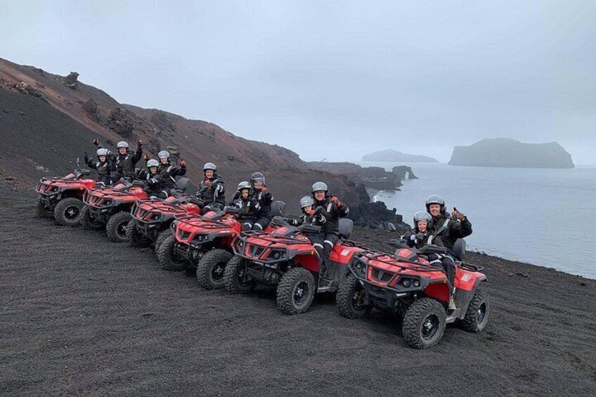 A group of people on red ATVs on a rocky beach with water and hills in the background. 2