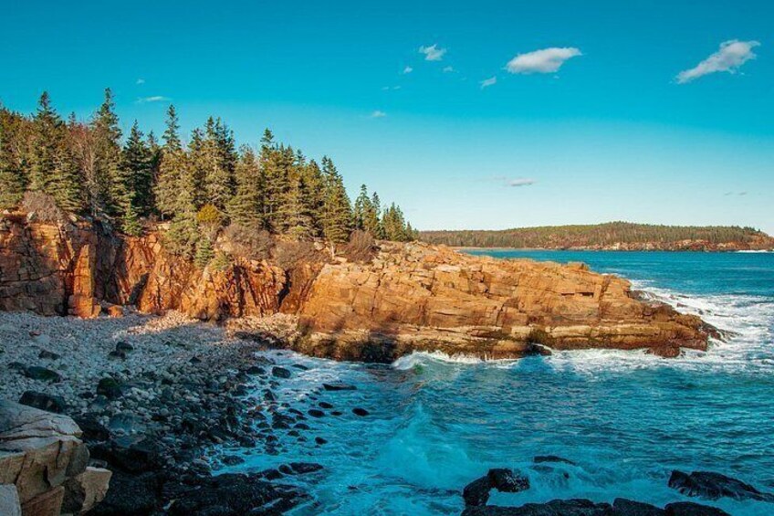 A rocky coastline with waves crashing against the shore, dense forest, and a clear blue sky. 3