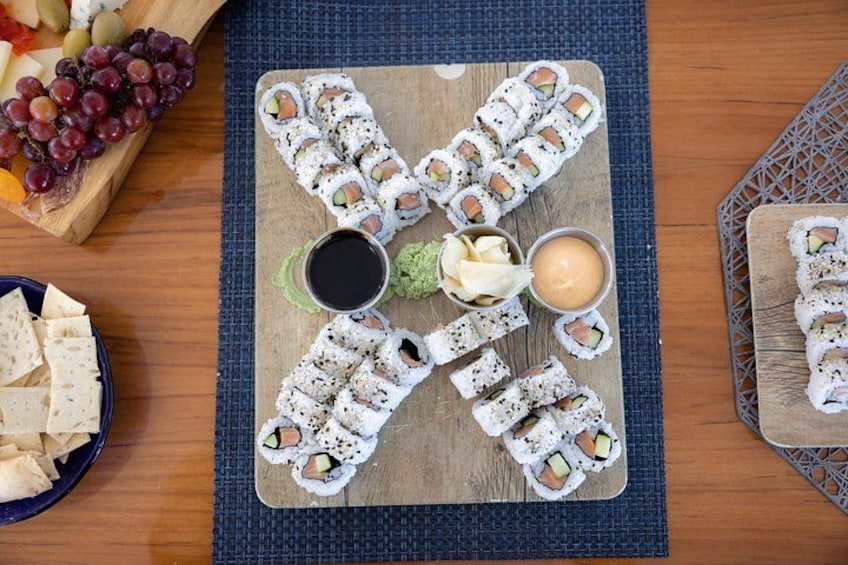 Un tagliere di legno con involtini di sushi disposti a croce, accompagnati da una ciotola di uva, un piatto di cracker e due salse per intingere. 2