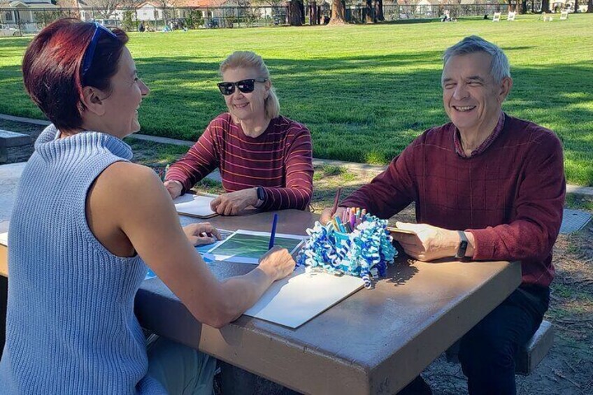 Three people sitting at a table in a park, engaged in an activity with papers and a blue object. 2