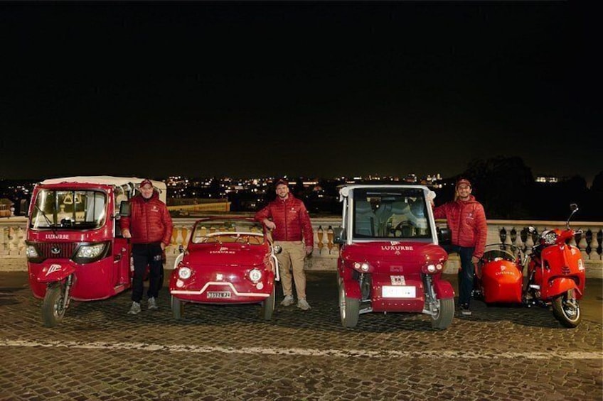 A group of people standing next to a red car and a red motorcycle on a cobblestone street at night. 2