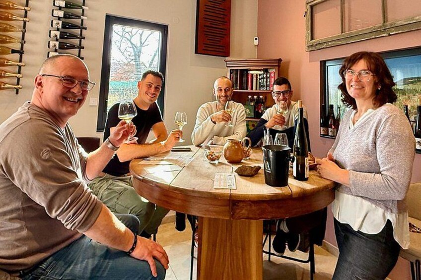 A group of people sitting around a round wooden table with wine glasses and bottles. 1