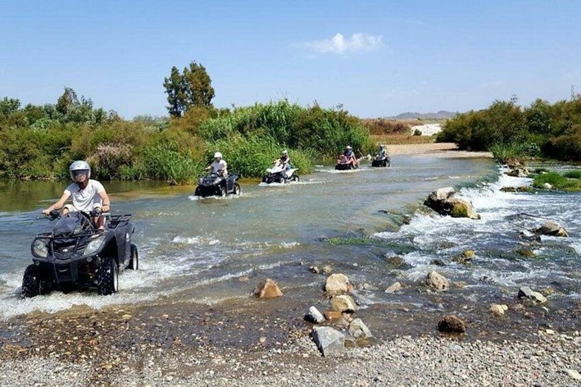 Des personnes conduisent des quads à travers une rivière peu profonde, dont les berges sont parsemées de rochers et de végétation. 1