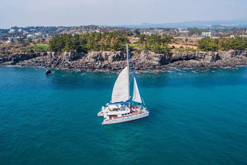 A sailboat on a turquoise sea with a rocky coastline and buildings in the background. 1