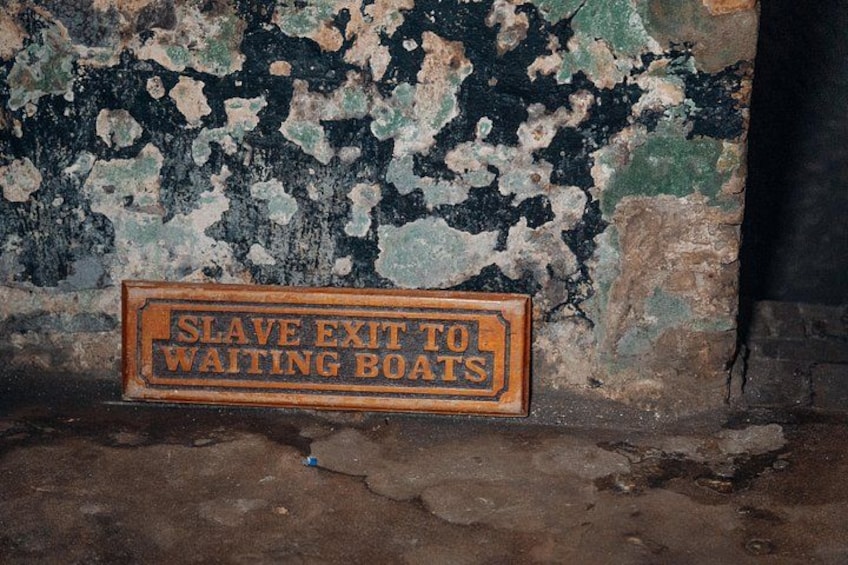 Ein verwittertes Schild mit abblätternder Farbe, auf dem 'SLAVE EXIT TO WAITING BOATS' steht, vor einer baufälligen Wand. 3