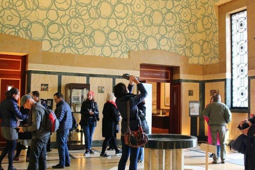 Un ambiente interno pubblico con pareti decorate, un tavolo di marmo circolare e persone impegnate in diverse attività. 2