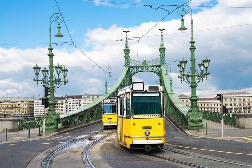 A yellow tram on a green bridge with street lamps and buildings in the background. 1