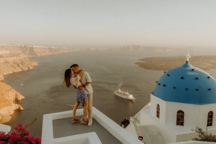 A couple embracing on a white building with a blue dome, overlooking a body of water with a ship in the distance. 4