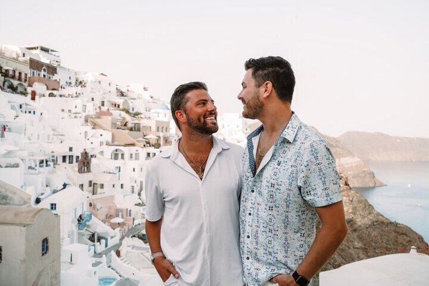Two men standing on a rooftop overlooking a white-washed coastal town with buildings and the sea in the background. 5