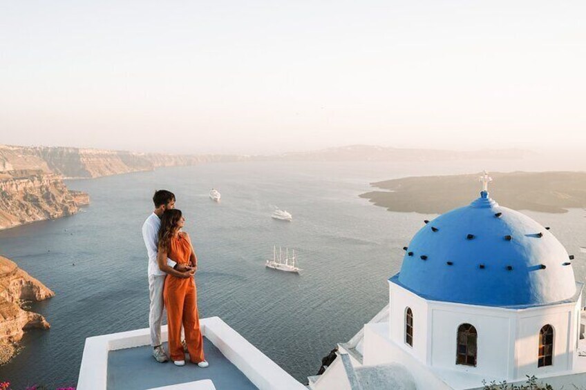A couple standing on a cliff overlooking a body of water with a blue-domed building in the foreground. 1