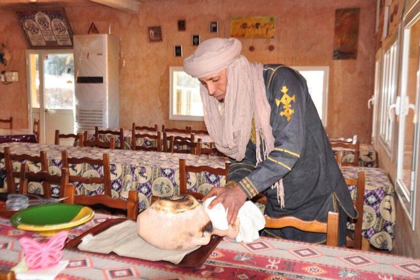 A person in traditional attire preparing food in a room with patterned tablecloths and wooden chairs. 4