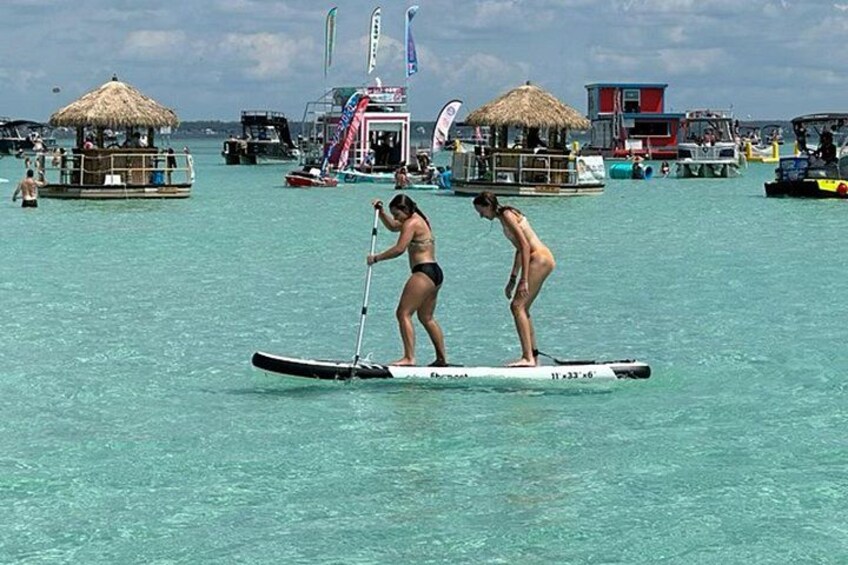 Two people paddleboarding on turquoise water with boats and thatched-roof structures in the background. 3