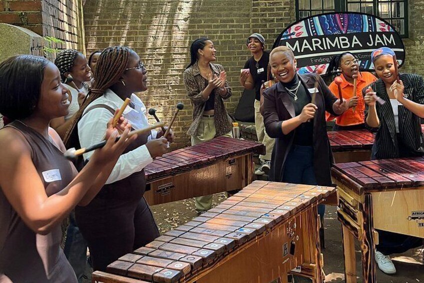 Un groupe de personnes joue de la marimba dans un espace extérieur, avec un mur de briques et une pancarte sur laquelle on peut lire « MARIMBA JAM ». 1