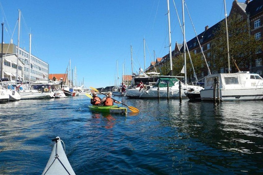 A calm waterway with boats, a person rowing a green kayak, and buildings along the shore. 2