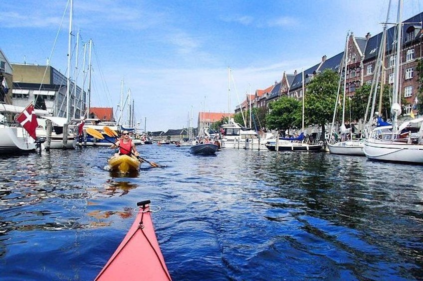 A canal with boats, a person in a kayak, and buildings along the water. 1