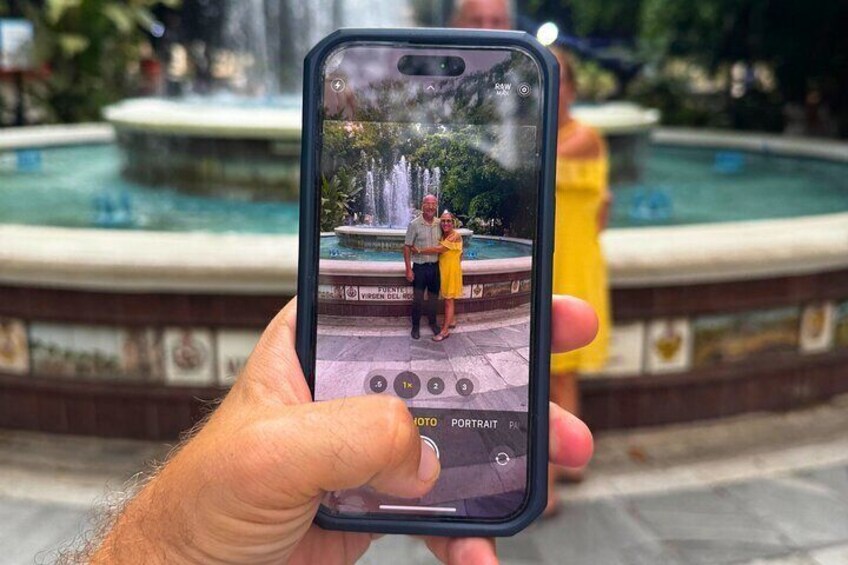 Una persona che tiene in mano uno smartphone con una foto di una fontana e due persone che si trovano di fronte. 2