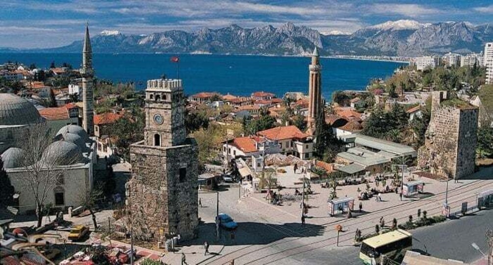 A coastal town with historic architecture, including a prominent tower and domed buildings, set against a backdrop of mountains and the sea. 1