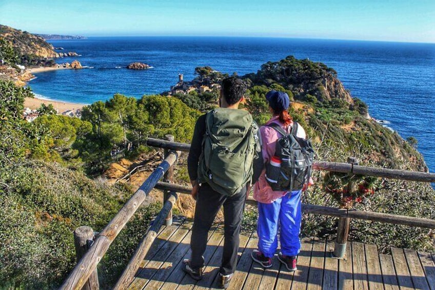 Dos personas de pie en un malecón de madera con vistas a un paisaje costero, una playa y acantilados rocosos. 1