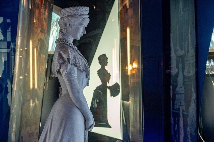 A statue of a woman in a white dress and lace collar, standing in a glass enclosure with a reflection of a building and a person in the background. 3