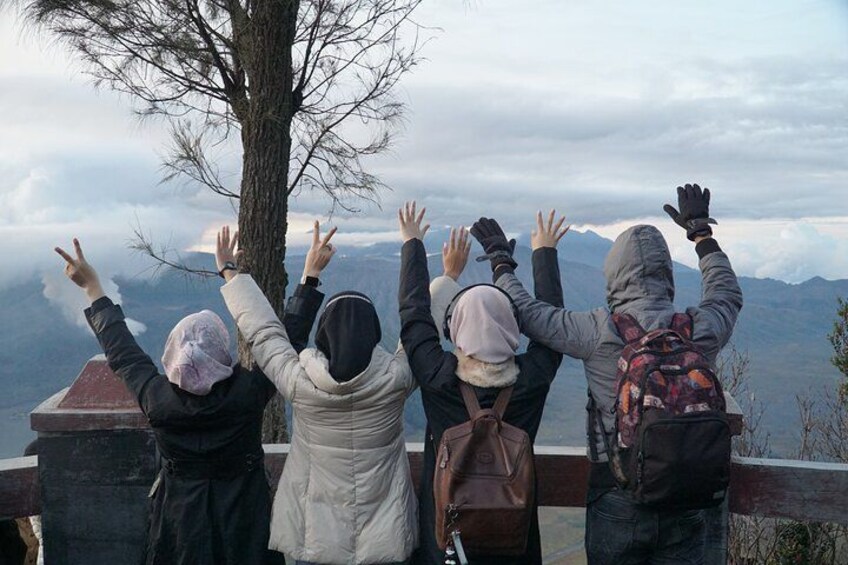 Vier Personen auf einer Aussichtsplattform mit erhobenen Händen, mit Blick auf eine gebirgige Landschaft. 2