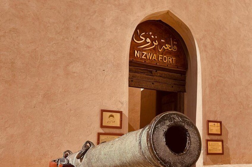Un cañón frente a un edificio con un letrero que dice 'NIZWA FORT'. 1