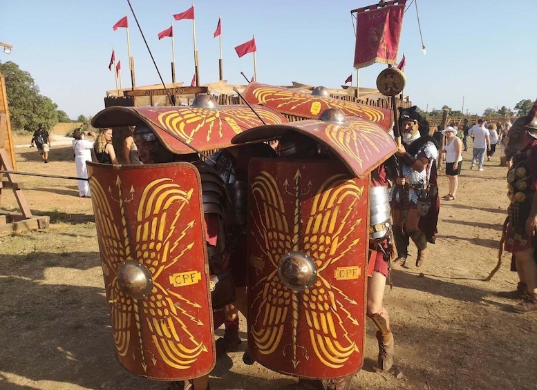 A group of people in Roman military armor with shields and helmets, standing in a line on a dirt ground. 4