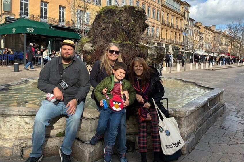 Une famille de quatre personnes est assise sur un banc de pierre, dans une place urbaine, avec des bâtiments en arrière-plan. 1