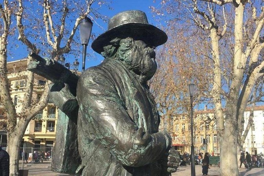 Une statue de bronze représentant un homme portant un chapeau et tenant une pipe, située dans un parc, avec des arbres et des bâtiments en arrière-plan. 3