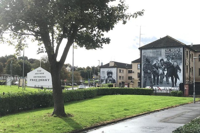 A grassy area with a sign, a tree, and a mural on a building. 1