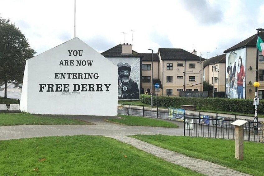 A large white sign with black lettering stating 'YOU ARE NOW ENTERING FREE DERRY' in front of a residential area with houses and a mural on a building. 3