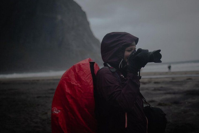 A person with a camera, wearing a red jacket, standing on a beach with a cliff in the background. 2