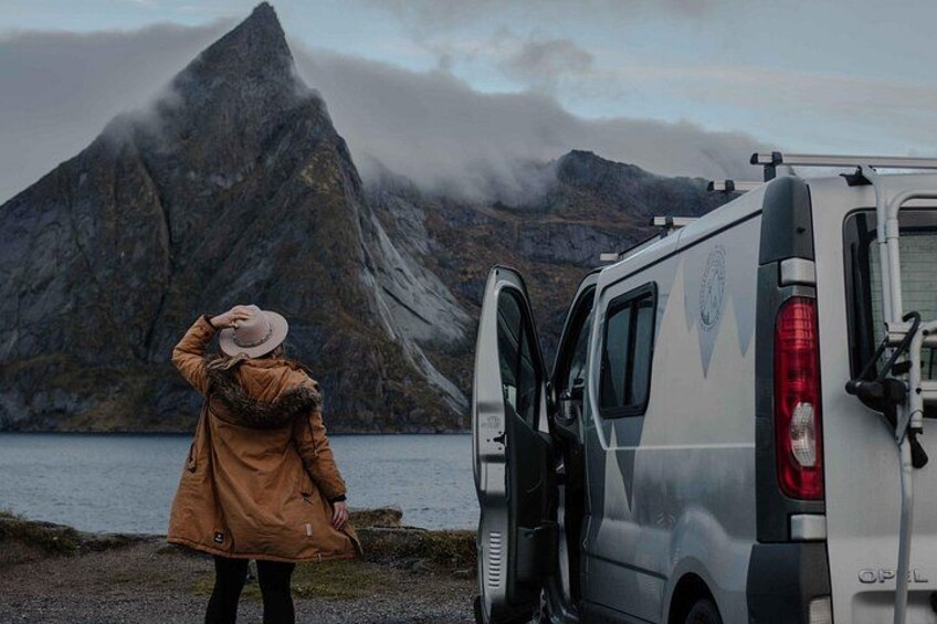 Eine Person in einem braunen Mantel und Hut steht neben einem weißen Van, im Hintergrund eine gebirgige Landschaft. 4