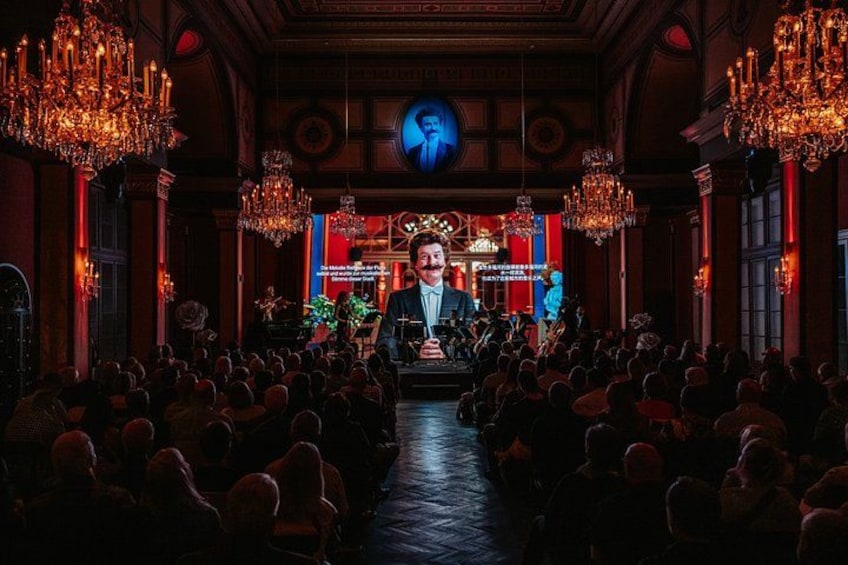 A theater interior with ornate chandeliers, a stage, and an audience seated. 3