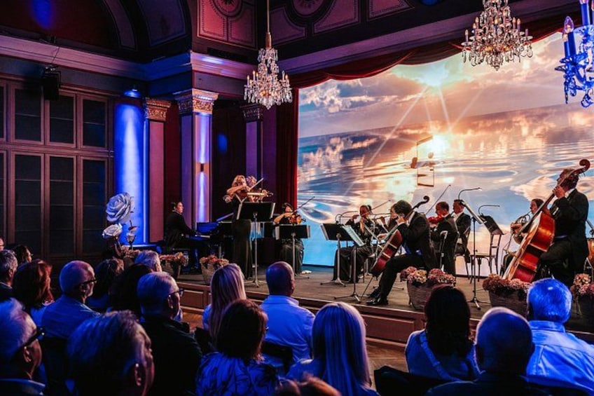 Ein Konzert klassischer Musik mit einem Orchester auf der Bühne, einem Pianisten und einem großen Publikum in einer Halle mit aufwendiger Architektur. 3