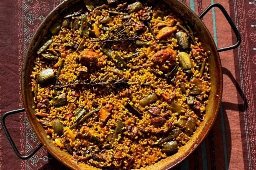 Een grote, ronde, ondiepe pan gevuld met een kleurrijk rijstgerecht, mogelijk paella, met zichtbare groenten en vlees. 5