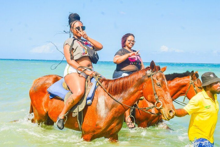 Zwei Personen reiten Pferde am Strand, das Wasser ist klar und der Himmel blau. 1