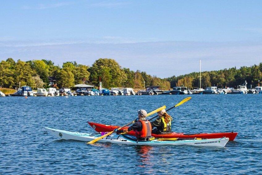 A person in a kayak on a calm body of water with a forested shoreline and several boats in the background. 3