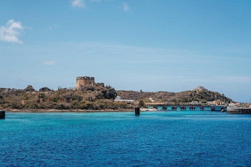 A coastal landscape with a historic fort on a hill, calm blue waters, and a pier extending into the sea. 3