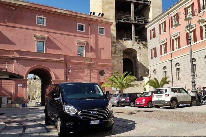 A black car parked on a cobblestone street, a pink building with an arched entrance, and a white building with red shutters. 1
