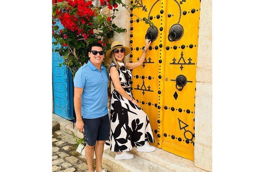 Un couple se tient devant une porte jaune ornée de motifs géométriques noirs, une porte bleue et, en arrière-plan, des fleurs rouges. 5