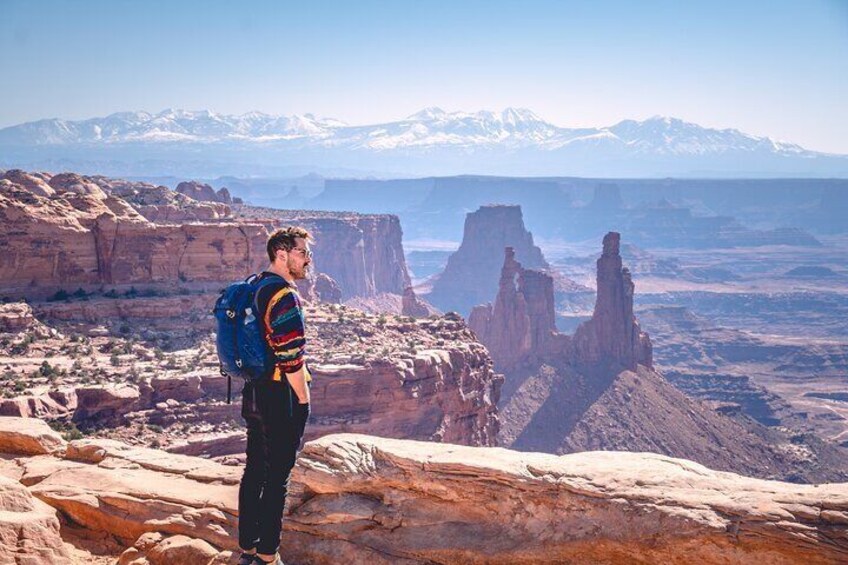 A person standing on a rocky outcrop overlooking a vast canyon with distinctive rock formations and snow-capped mountains in the distance. 5