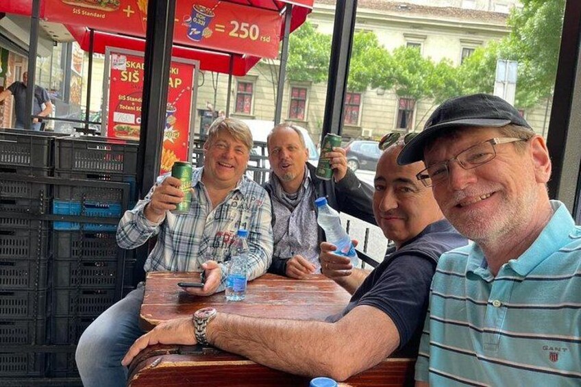 A group of people sitting at a table in an outdoor cafe with a red awning and a vending machine in the background. 2