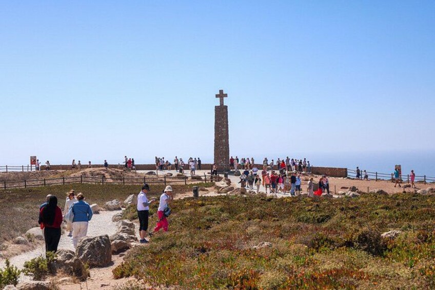 Zona costera con un monumento cruciforme destacado, terreno rocoso y una multitud de personas. 2