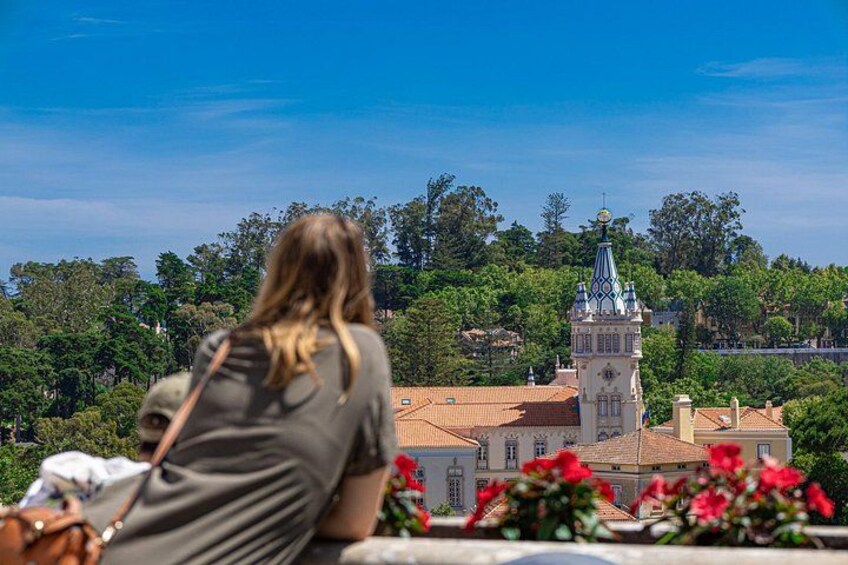 A woman sitting on a ledge overlooking a historic building with a tower, surrounded by lush greenery. 2