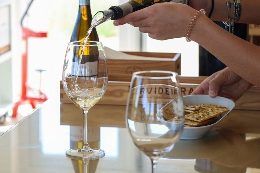 A person pouring wine into a glass, with a bottle and a bowl of crackers on the table. 2