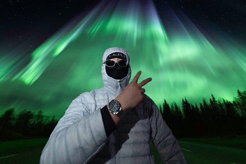 A person in a white puffer jacket and black mask posing with a peace sign against a backdrop of the Northern Lights. 2