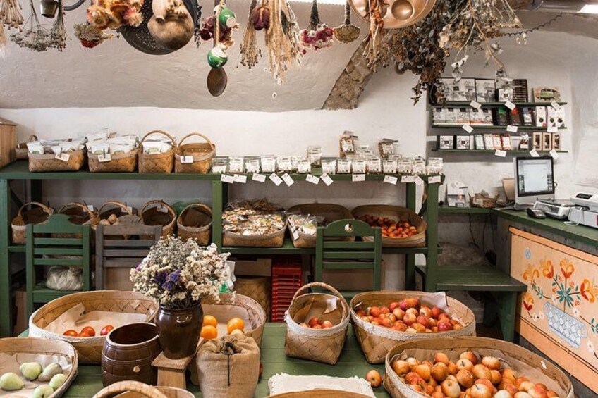 Un puesto de mercado con cestas de frutas, un jarrón con flores y estanterías llenas de productos. 5