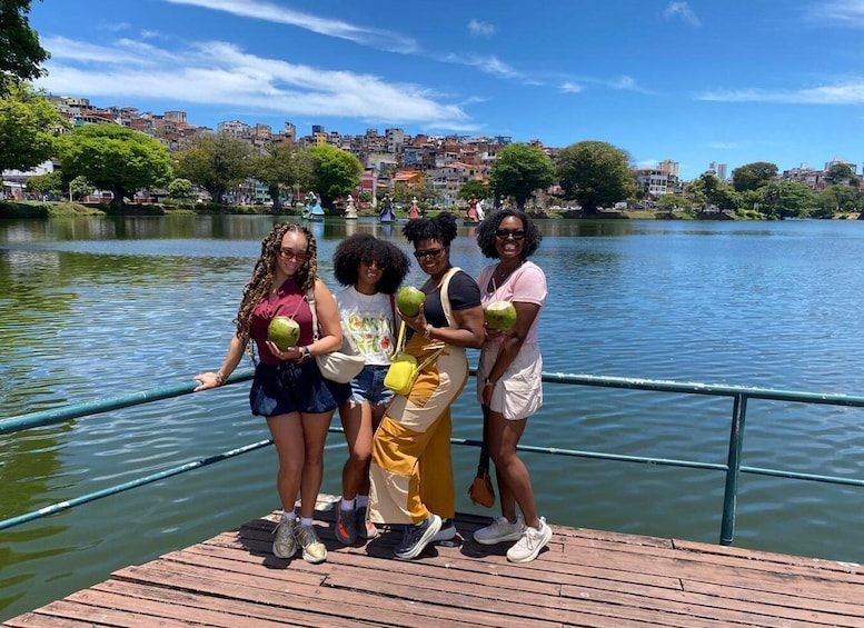 Un grupo de personas en un muelle de madera junto a un lago, con un paisaje urbano al fondo. 3