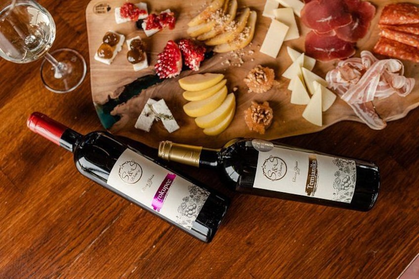 Dos botellas de vino, una tabla de madera con diversos quesos, embutidos y frutas, y una copa de vino blanco sobre una mesa de madera. 1