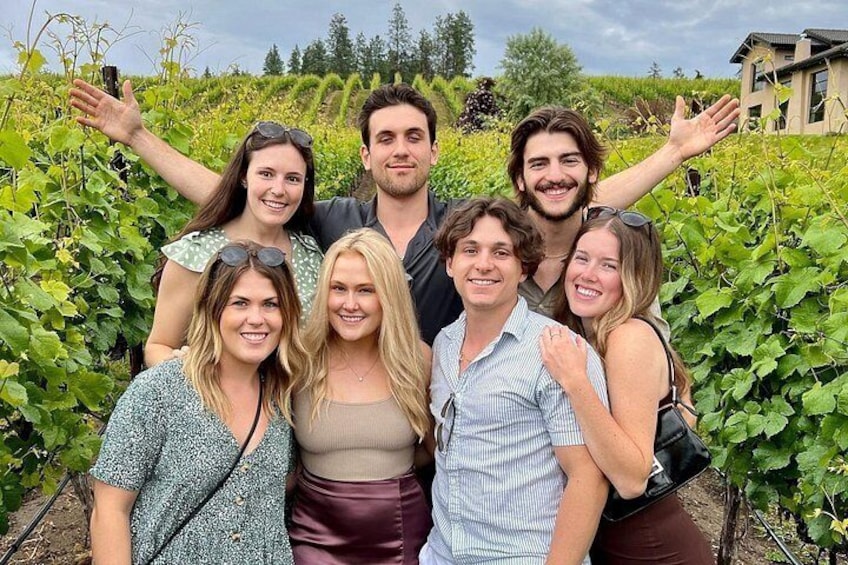 A group of people in a vineyard with rows of grapevines and a building in the background. 1